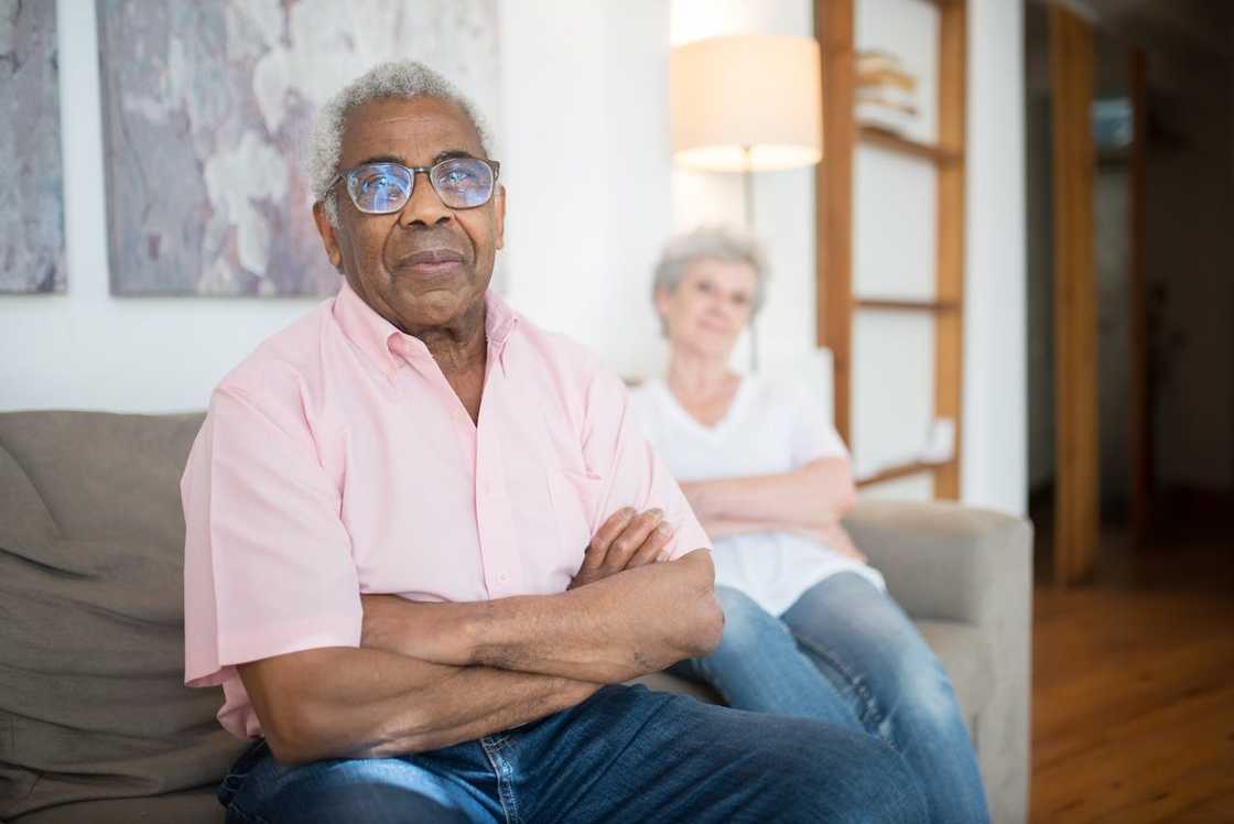 An elderly man sits on a couch with his arms crossed while a woman sits behind him. An elderly man sits on a couch with his arms crossed while a woman sits behind him.