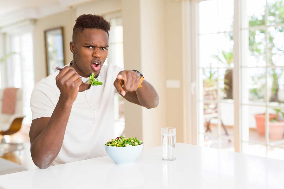 Person points toward the camera while holding a fork with salad in a bright dining area.