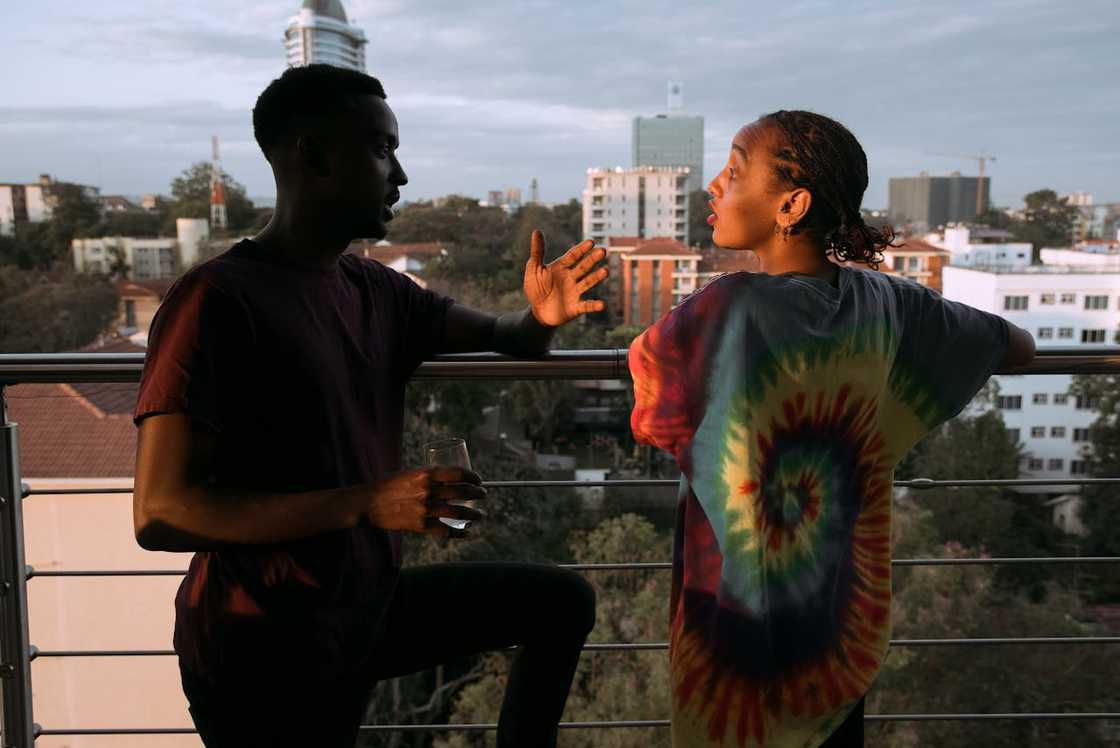 A man gestures while speaking to a woman on a balcony overlooking a city. A man gestures while speaking to a woman on a balcony overlooking a city.