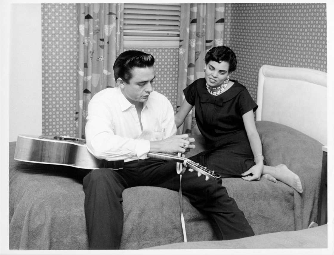 Johnny Cash sits on a bed tying his guitar strap as his first wife Vivian Liberto