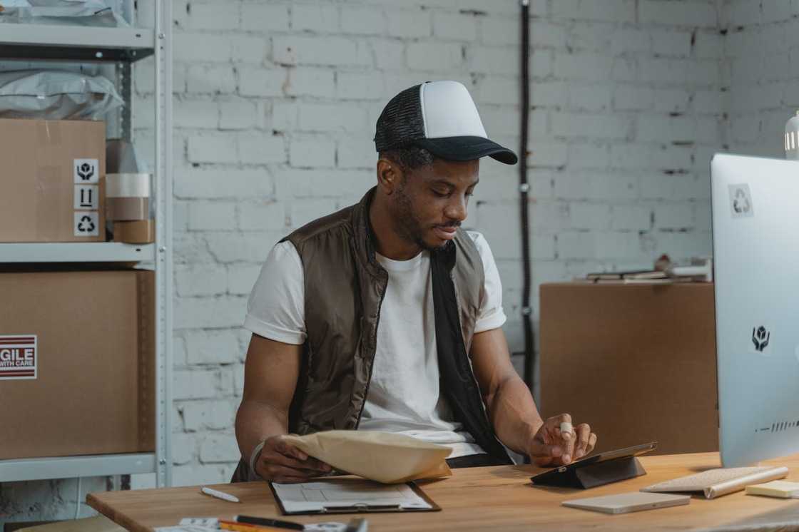 A warehouse worker seated at a desk, checking a package and using a tablet. A warehouse worker seated at a desk, checking a package and using a tablet.