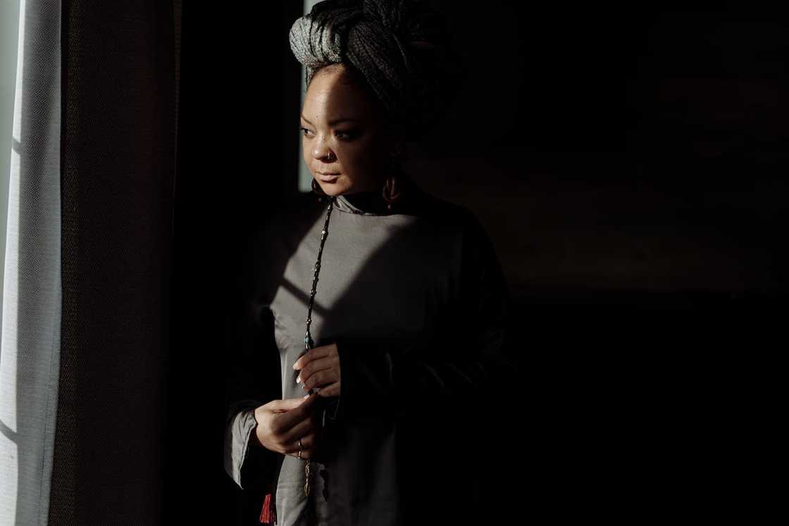 A woman standing by a window in low light, looking thoughtful.