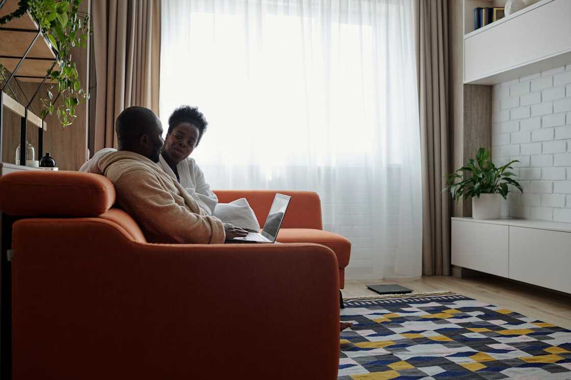 A couple sits together on an orange sofa, talking while one uses a laptop. A couple sits together on an orange sofa, talking while one uses a laptop.