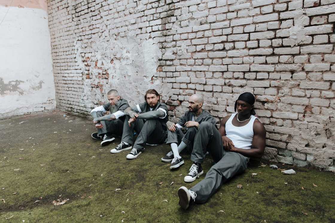 A group of inmates sit against a brick prison wall.