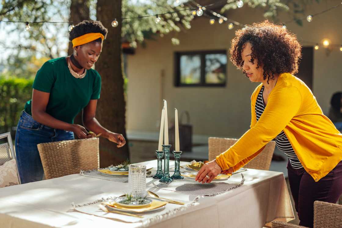 Two people set an outdoor dining table with white linens, candles, and rosemary sprigs. Two people set an outdoor dining table with white linens, candles, and rosemary sprigs.