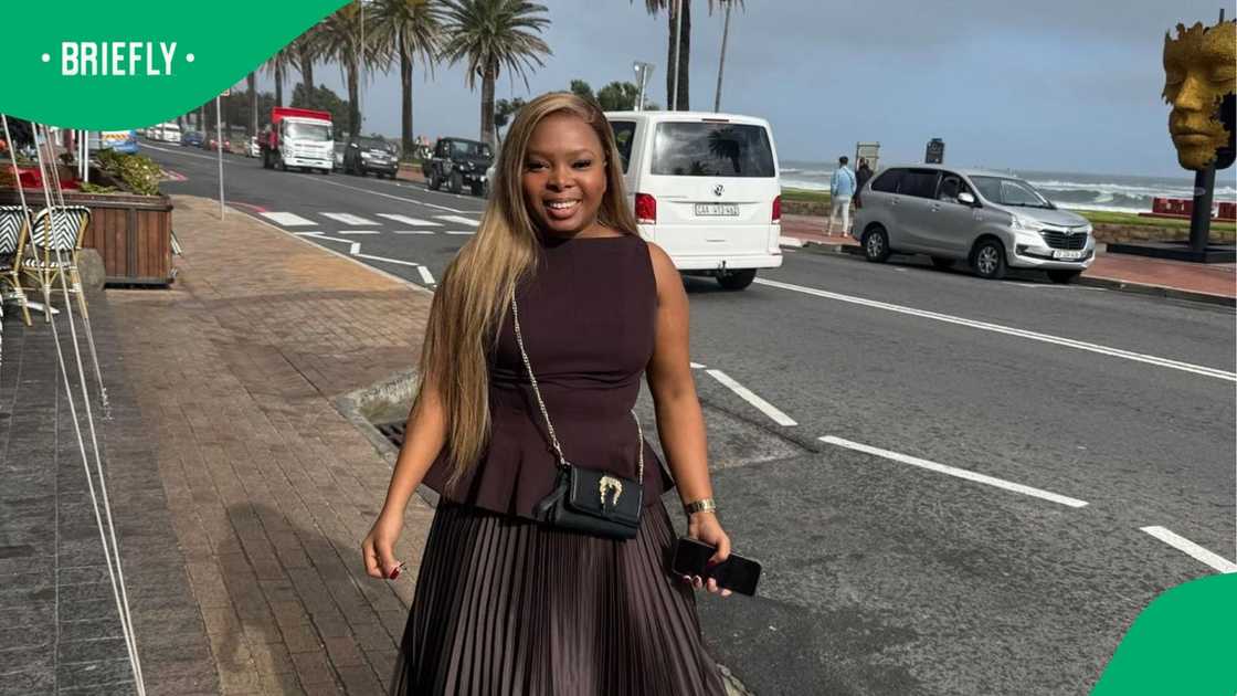 A young woman with long blonde hair, wearing a dark brown peplum top and pleated skirt, smiles at the camera while standing on a sidewalk next to a road with palm trees and the ocean in the background. A young woman with long blonde hair, wearing a dark brown peplum top and pleated skirt, smiles at the camera while standing on a sidewalk next to a road with palm trees and the ocean in the background.