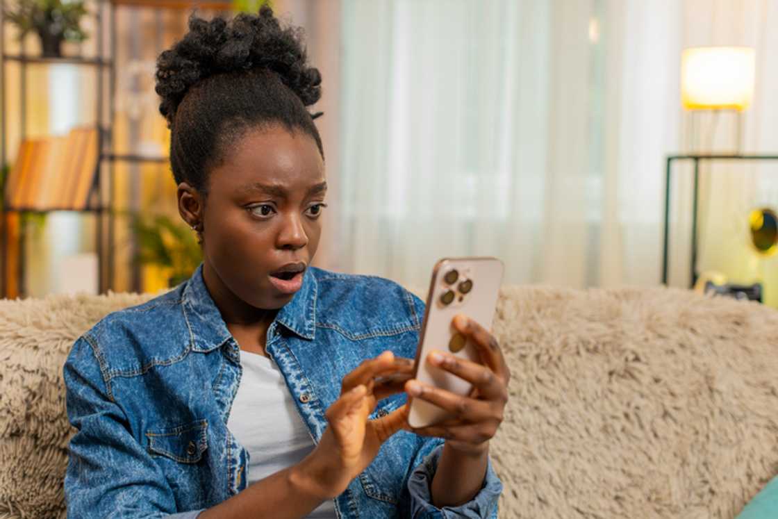 A woman looking at her phone, shocked, while sitting on a couch.