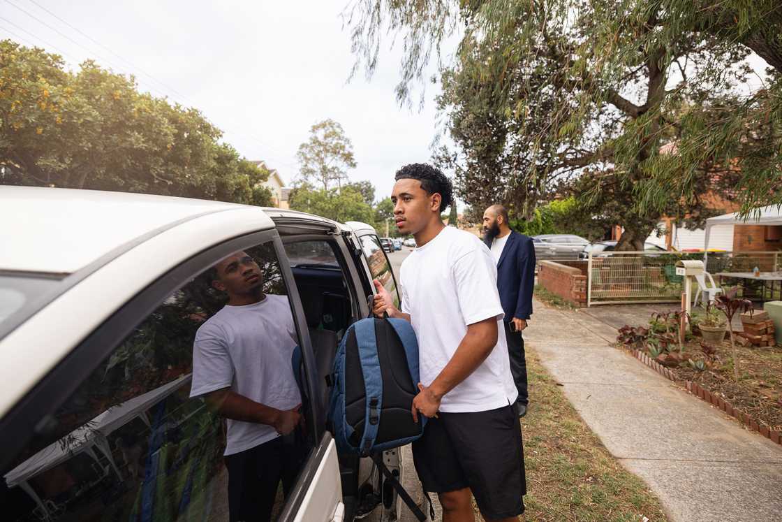 A young man loads a backpack into a car while an older man watches from the driveway.