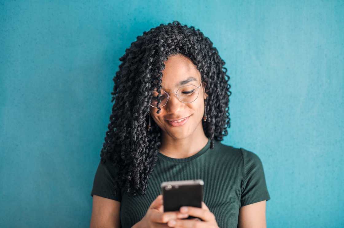 A woman stands against a blue wall smiling as she looks at her phone.