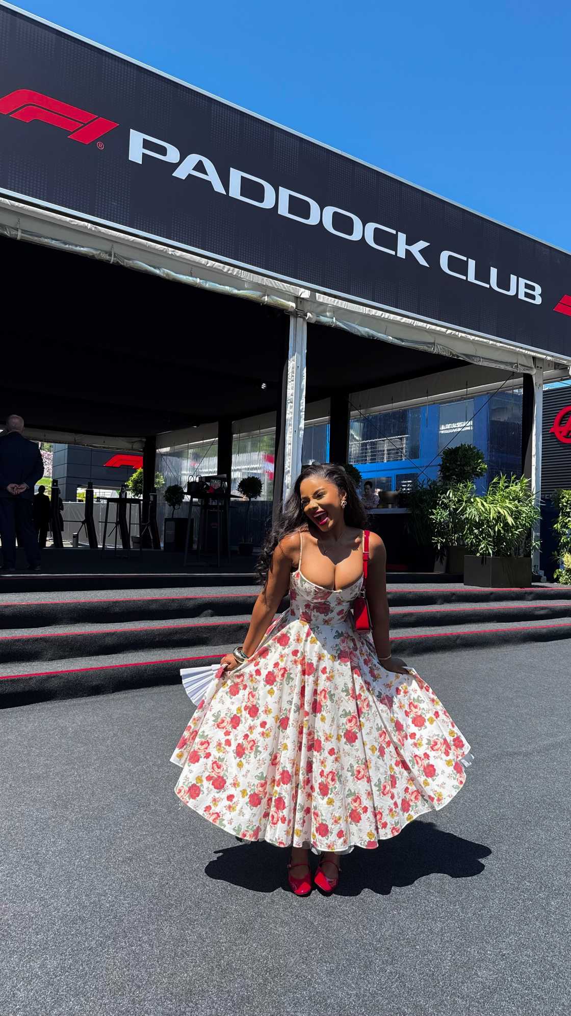 Pamela Mtanga twirls in a red and cream floral dress outside the Paddock Club entrance at the Austrian Grand Prix.