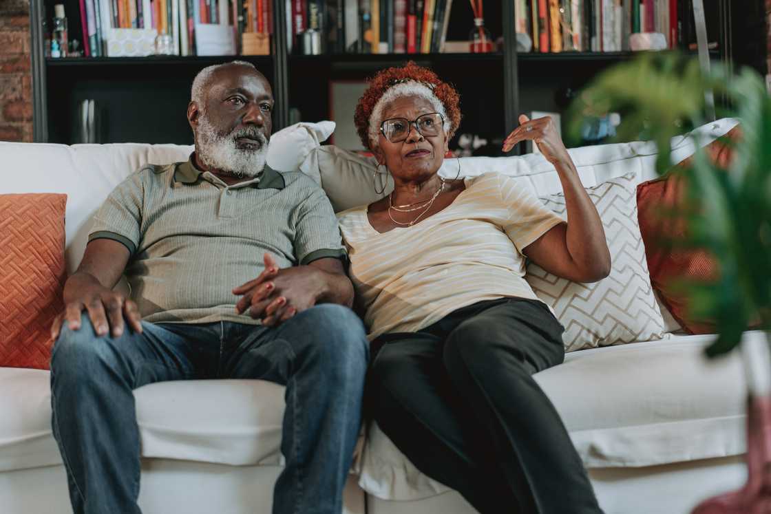 Two older adults sit on a white couch holding hands.