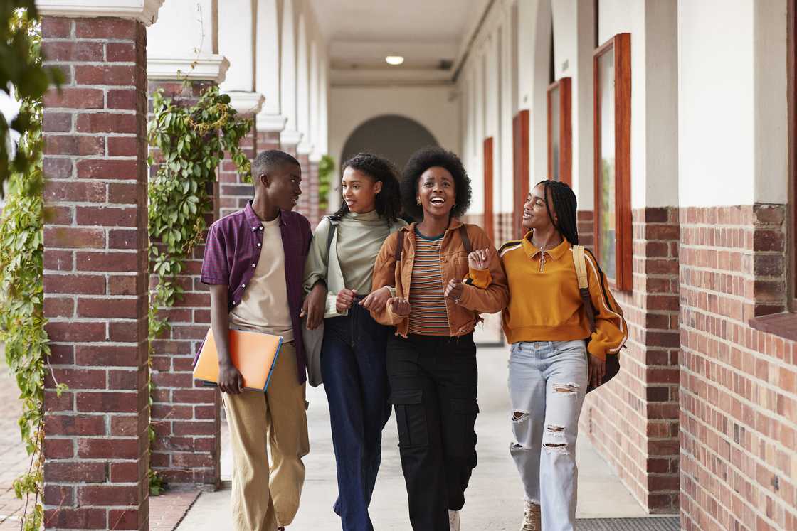 Happy friends talking while walking with arms in arms at high school corridor.