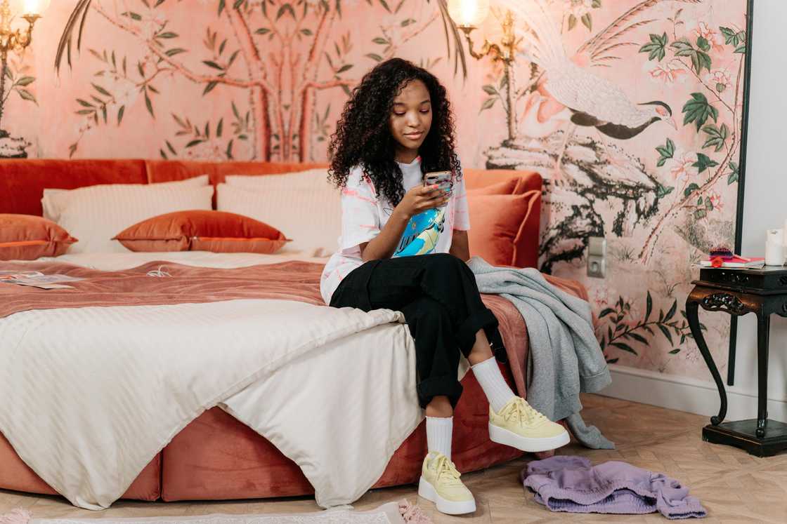 A woman sitting on the bed with crossed legs uses a smartphone