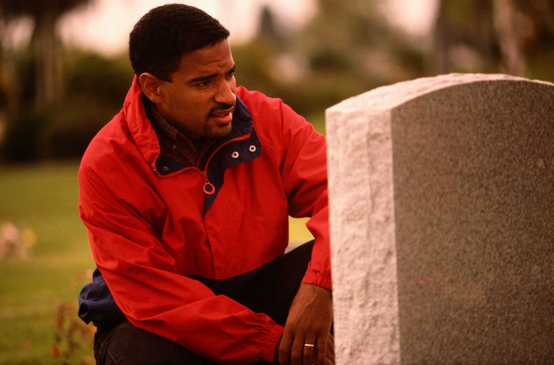 Person in red and navy jacket kneels beside a grey gravestone. Person in red and navy jacket kneels beside a grey gravestone.