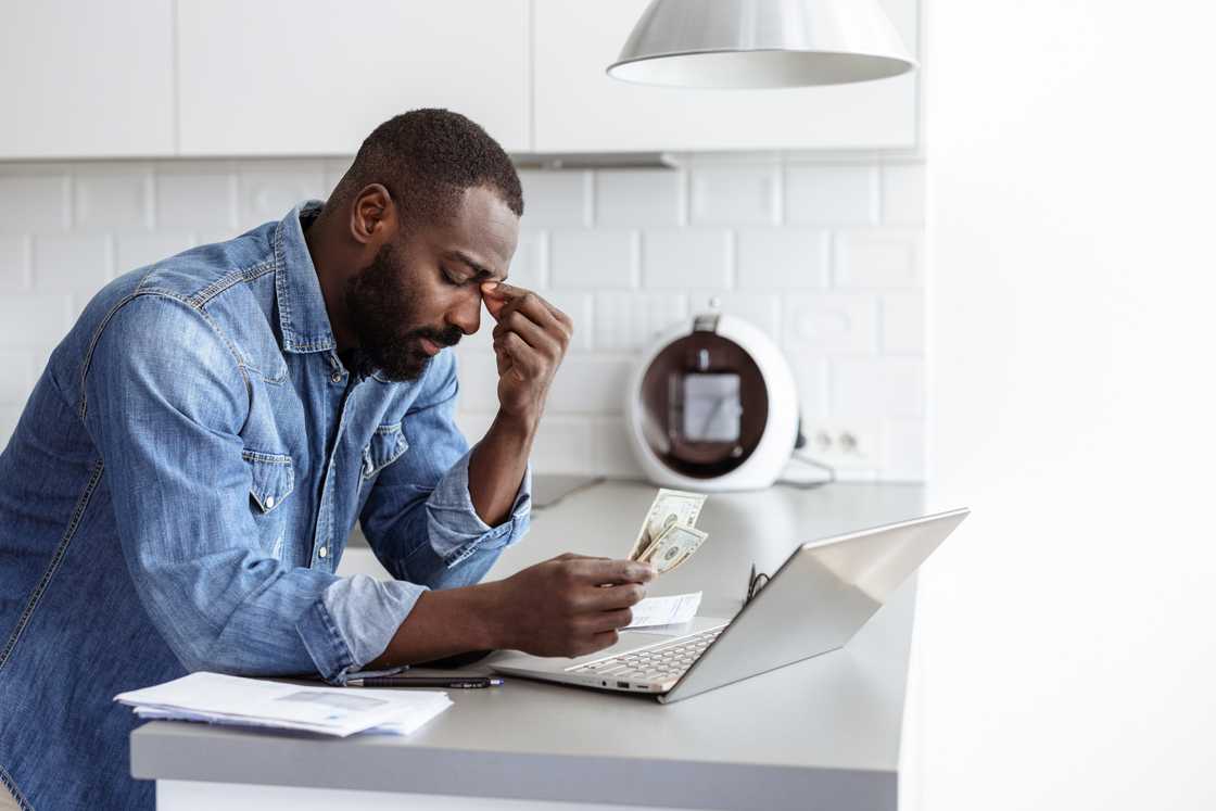 Young man in a casual setting looks concerned while reviewing finances, holding cash and using a laptop. This scene reflects financial stress, budget challenges, and personal finance management