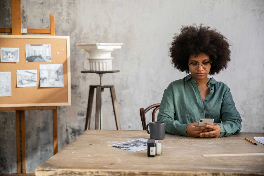 A woman in an afro hairstyle and a green shirt is using a smartphone on a desk