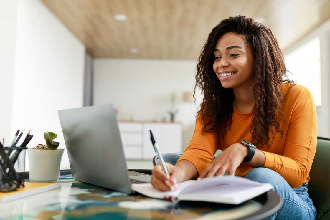 A woman sitting at a desk working on a laptop and writing in paper notebook.