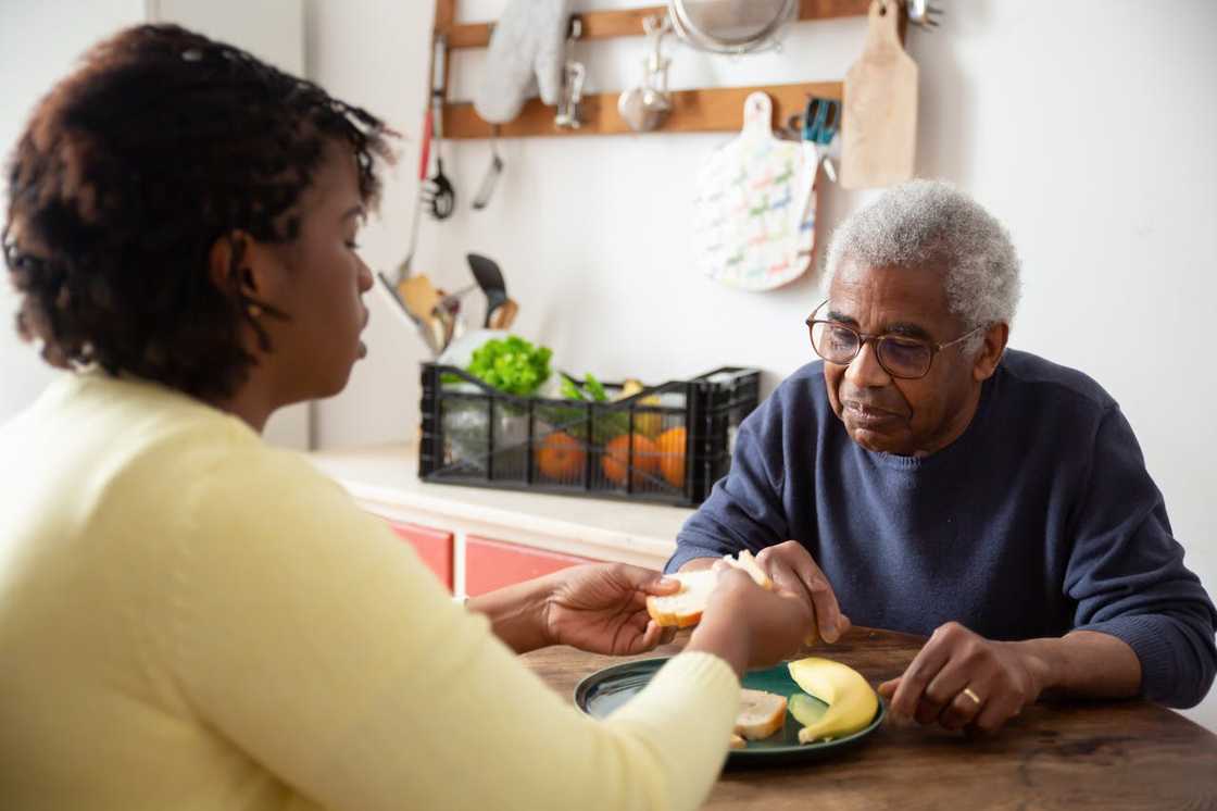 Woman handing food to elderly man at kitchen table. Woman handing food to elderly man at kitchen table.