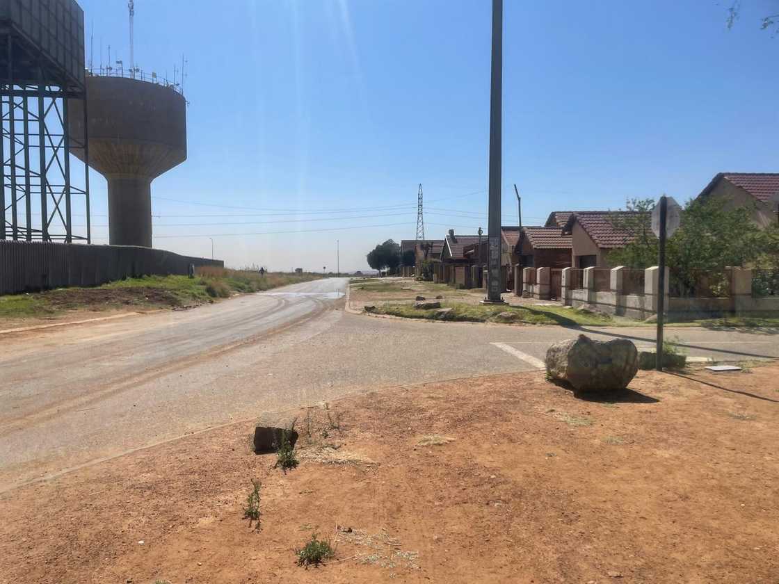 A wide shot of a dusty suburban street with a large water tower on the left and a row of houses on the right. A wide shot of a dusty suburban street with a large water tower on the left and a row of houses on the right.