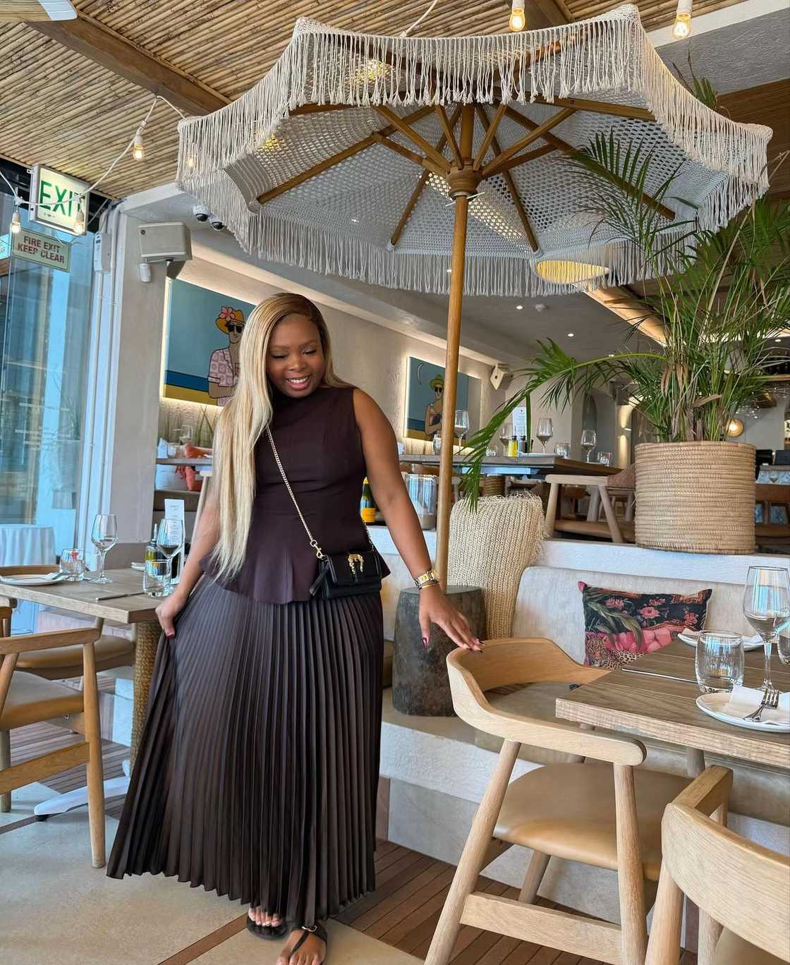 A young woman with long blonde hair, wearing a dark brown sleeveless top and matching pleated skirt, smiles while standing next to a wooden chair in a stylish restaurant decorated with plants and a fringed umbrella-style light. A young woman with long blonde hair, wearing a dark brown sleeveless top and matching pleated skirt, smiles while standing next to a wooden chair in a stylish restaurant decorated with plants and a fringed umbrella-style light.