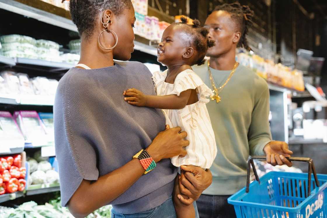 An adult holds a toddler while another adult carries a shopping basket. An adult holds a toddler while another adult carries a shopping basket.