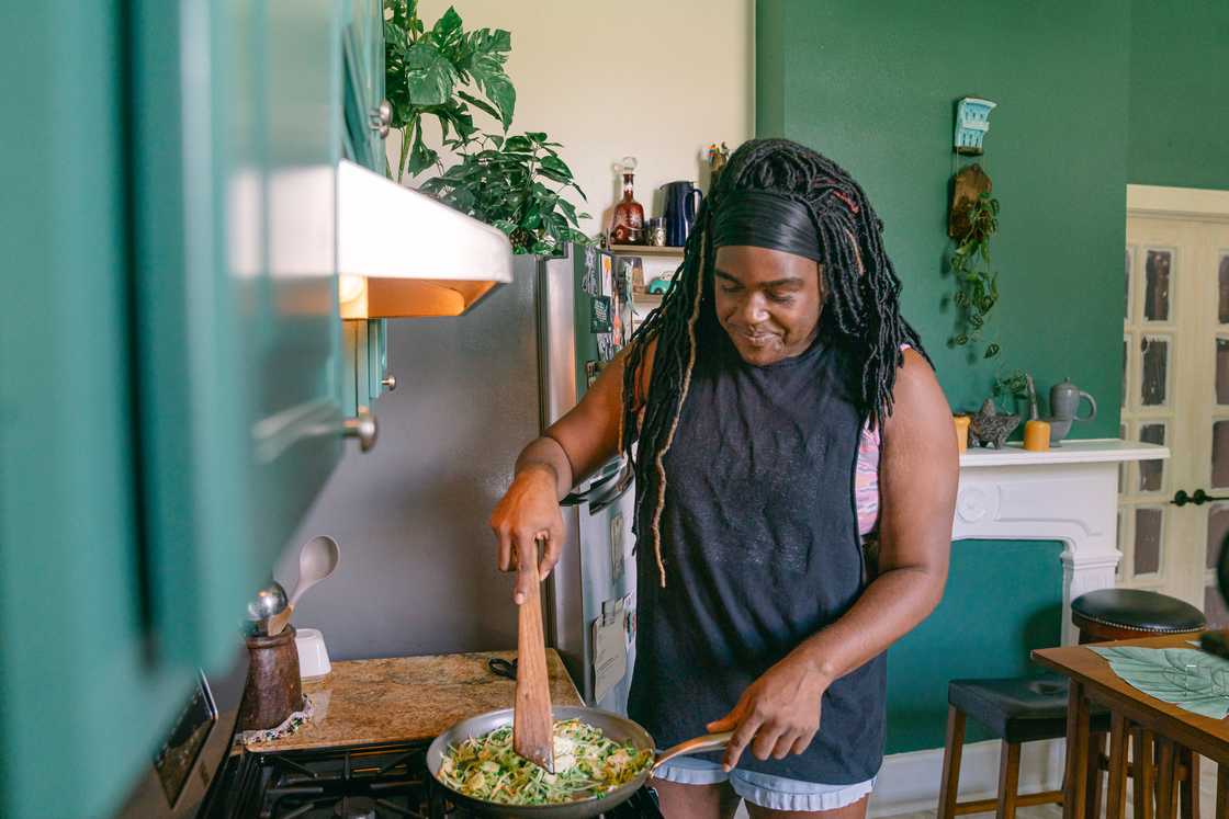 A person stirs food in a pan in a green kitchen filled with plants. A person stirs food in a pan in a green kitchen filled with plants.