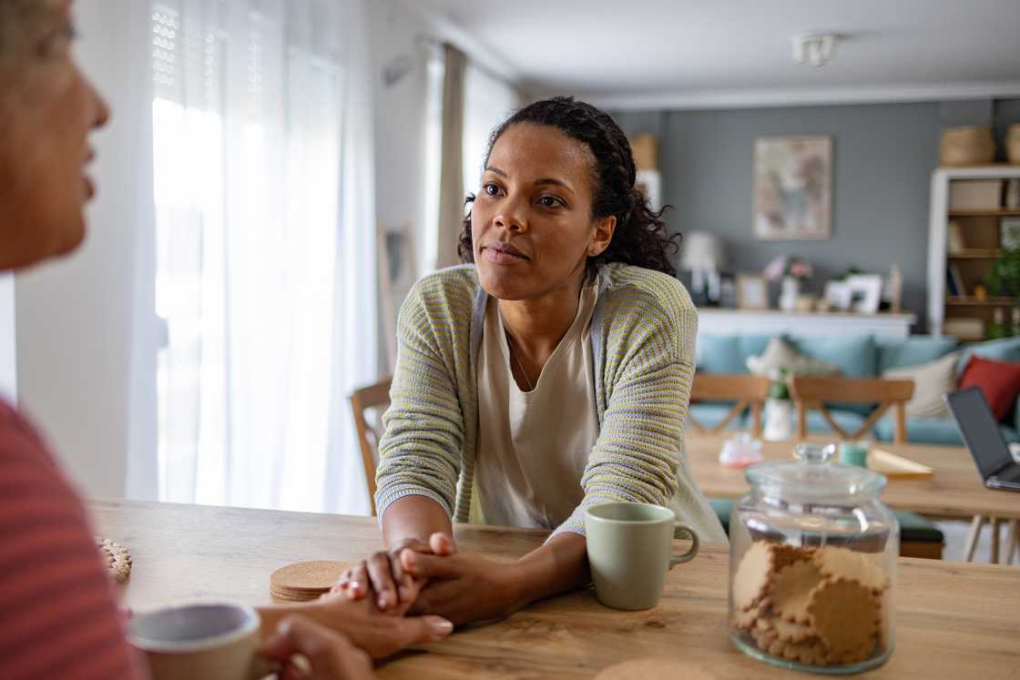 Two people sit at a wooden table; one speaks while the other listens and holds their hand.