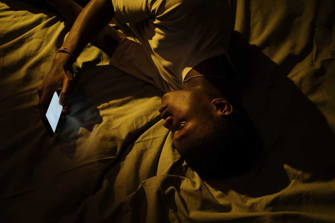A man lies on a bed holding a phone in dim light. A man lies on a bed holding a phone in dim light.