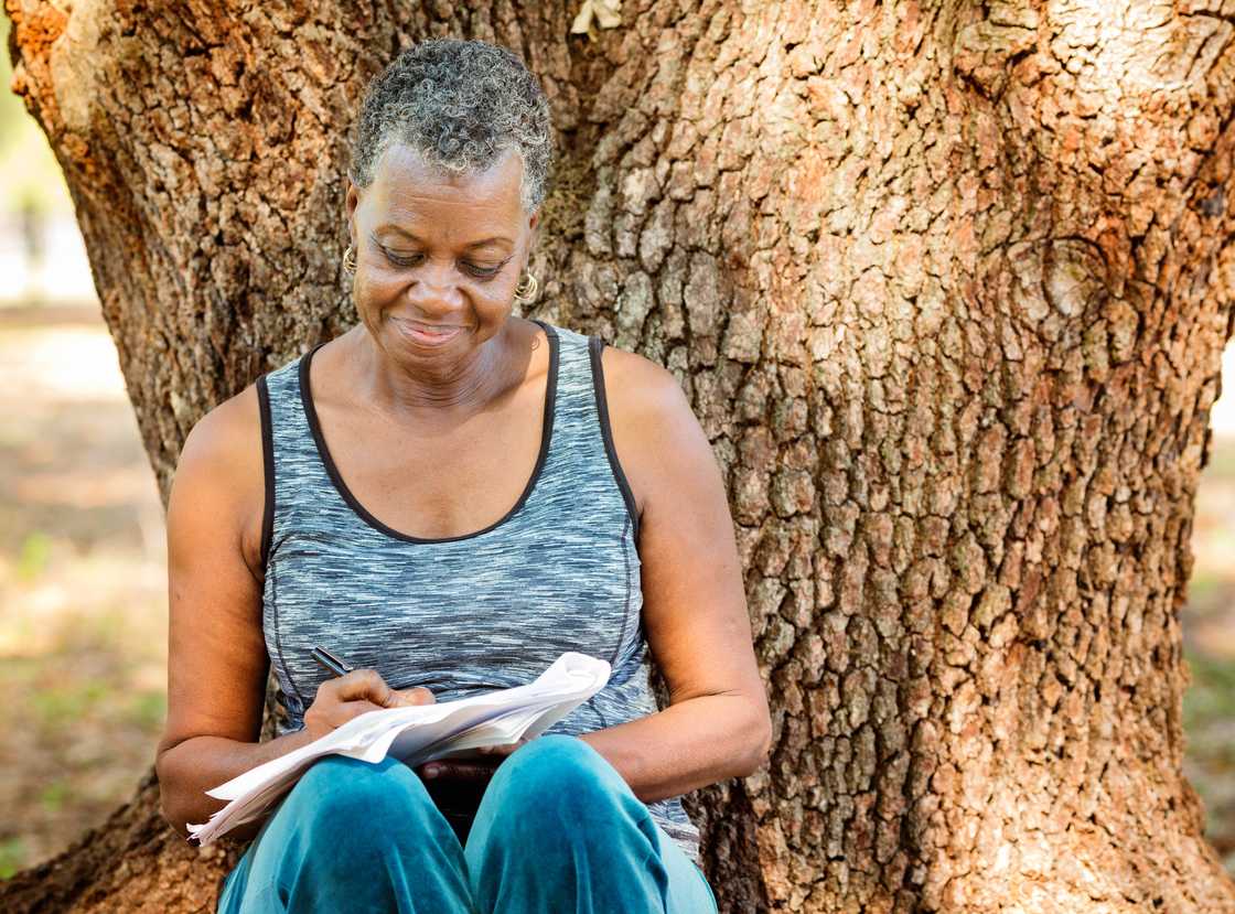 Older adult writes in a notebook while seated against a large tree outdoors. Older adult writes in a notebook while seated against a large tree outdoors.