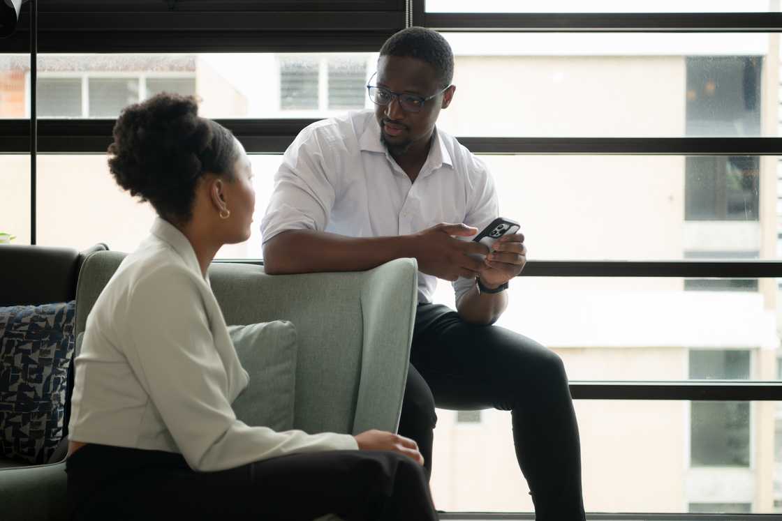 Two people sit on a couch, one showing something on a phone.