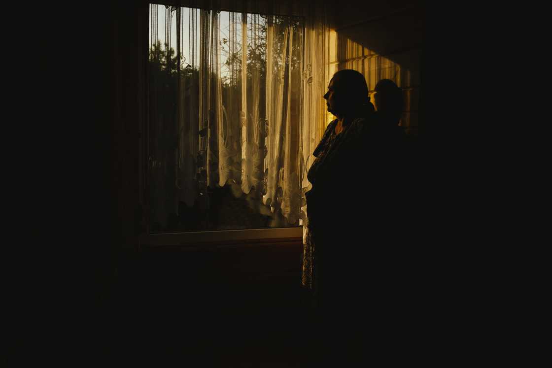 Silhouetted person stands near a sunlit window in a dimly lit room.