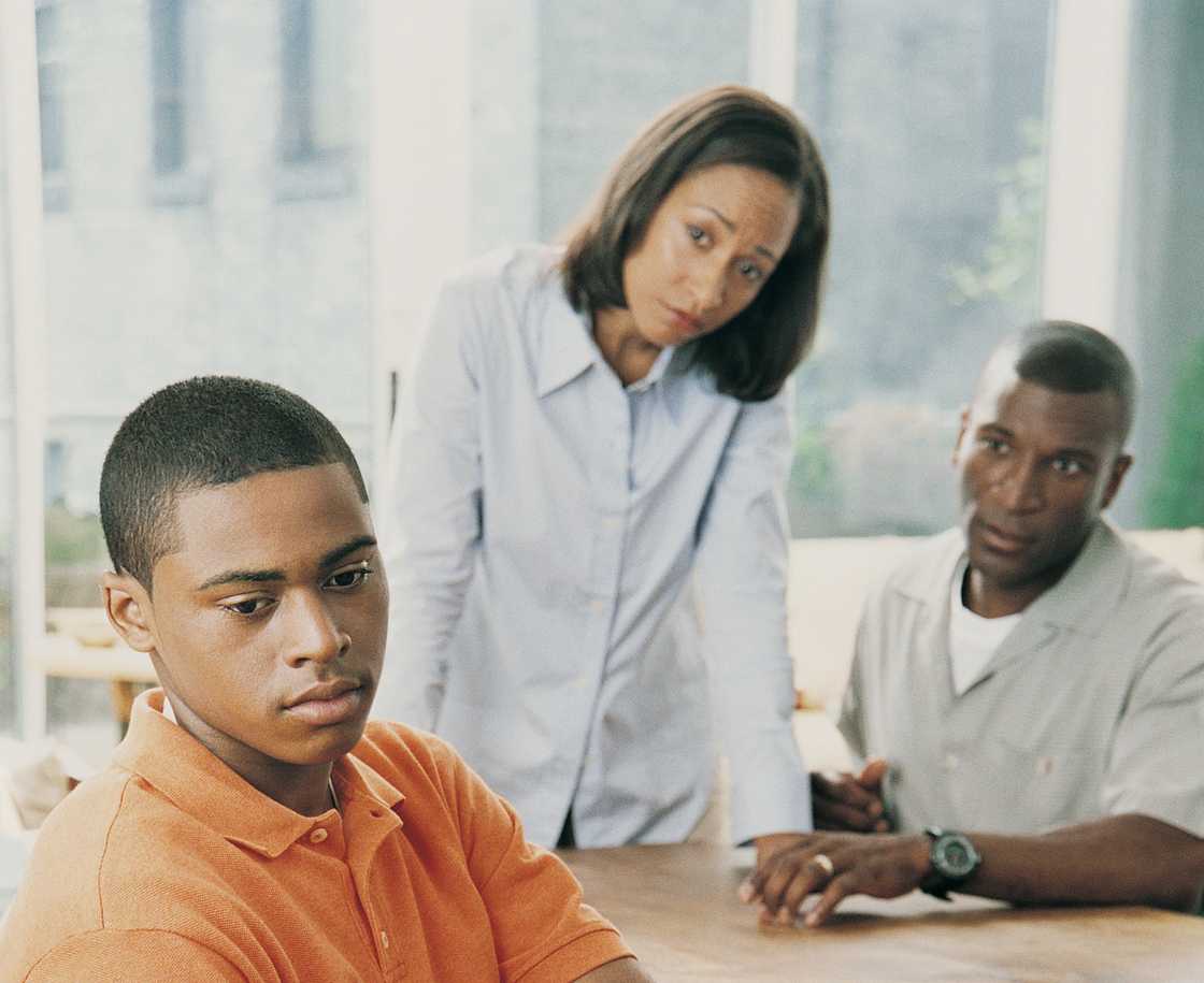 Young person sits quietly while two adults look on with concern. Young person sits quietly while two adults look on with concern.