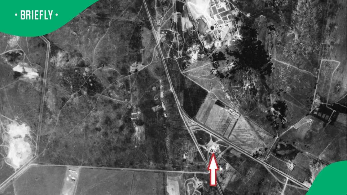 A wider black-and-white aerial photograph captured an expanded landscape of roads, highway interchanges, and residential plots, with a blue arrow pointing to a location at the bottom of the frame A wider black-and-white aerial photograph captured an expanded landscape of roads, highway interchanges, and residential plots, with a blue arrow pointing to a location at the bottom of the frame