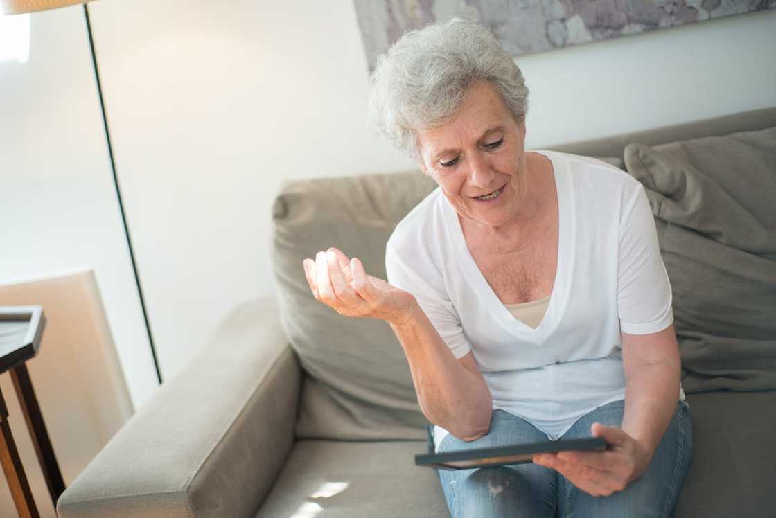 An elderly woman sitting on a sofa holding a portrait, gesturing in confusion.