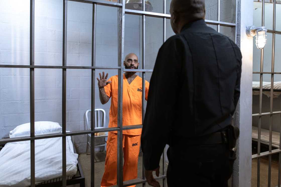 A man in prison uniform standing behind bars, speaking to a correctional officer. A man in prison uniform standing behind bars, speaking to a correctional officer.