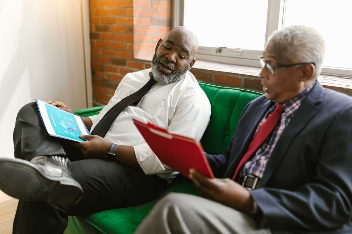 Two businessmen sit on a sofa discussing documents during a meeting.