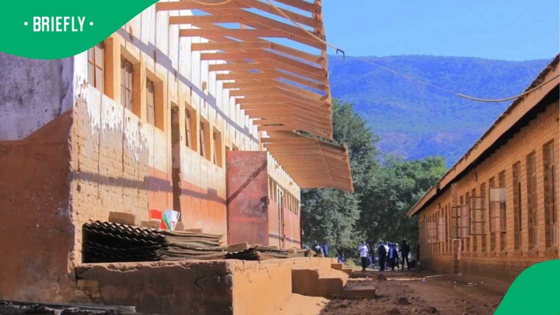 The weathered exterior of the dilapidated school facility showed broken brickwork, unpaved walkways, and a missing roof structure against a mountain backdrop The weathered exterior of the dilapidated school facility showed broken brickwork, unpaved walkways, and a missing roof structure against a mountain backdrop