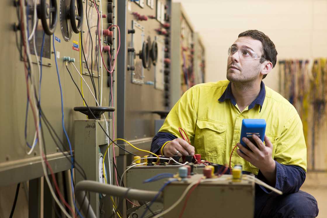 A stock photo of an electrician wearing yellow safely jacket testing an electrical switchboard