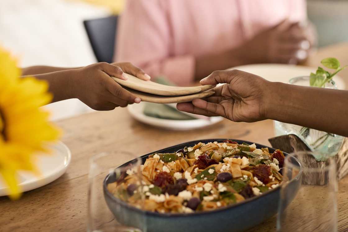 Two people pass stacked wooden plates across a dining table set with pasta salad and a sunflower. Two people pass stacked wooden plates across a dining table set with pasta salad and a sunflower.