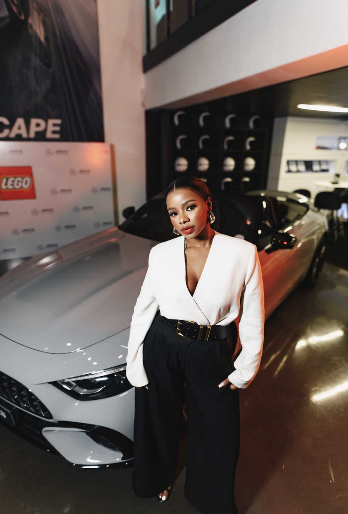 Pamela Mtanga poses in a white blazer, black wide-leg trousers and a statement belt in front of a Mercedes-AMG showroom display.