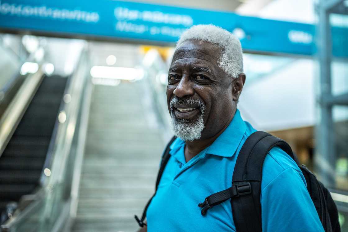 Older person with grey hair and beard smiles while standing in front of an escalator. Older person with grey hair and beard smiles while standing in front of an escalator.