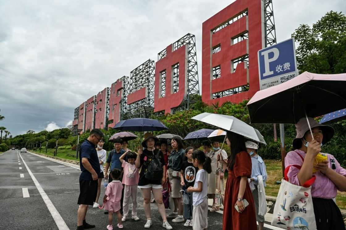 People in Xiamen wait for a bus on a street next to a sign that reads in Chinese "One country, two systems: reunify China" People in Xiamen wait for a bus on a street next to a sign that reads in Chinese "One country, two systems: reunify China"
