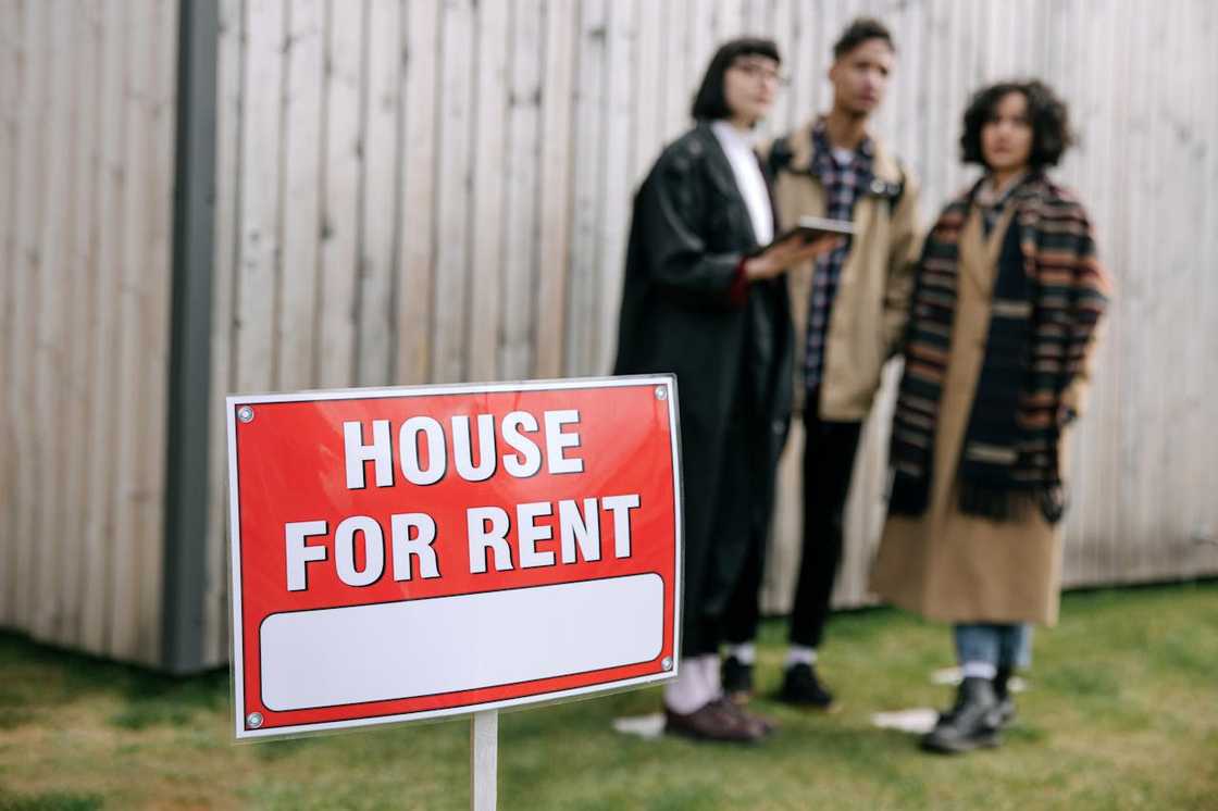 A group of people stand behind a house for rent sign. A group of people stand behind a house for rent sign.