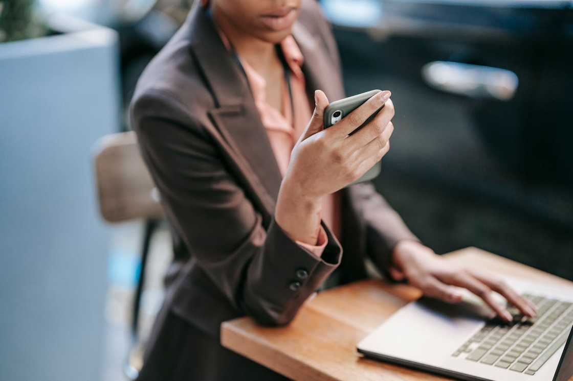 A woman sits at a desk using a phone beside an open laptop.