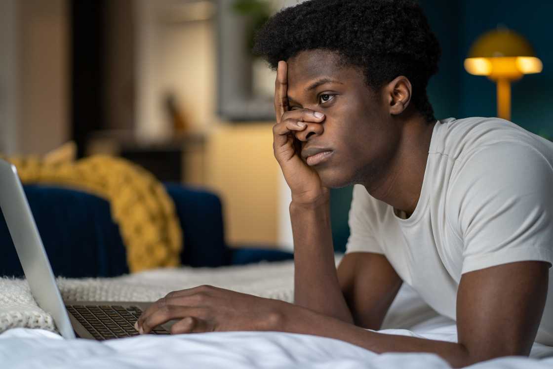 Frustrated, lazy black man on bed with laptop