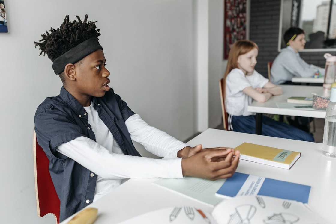 A student sits at a desk while classmates work behind him. A student sits at a desk while classmates work behind him.