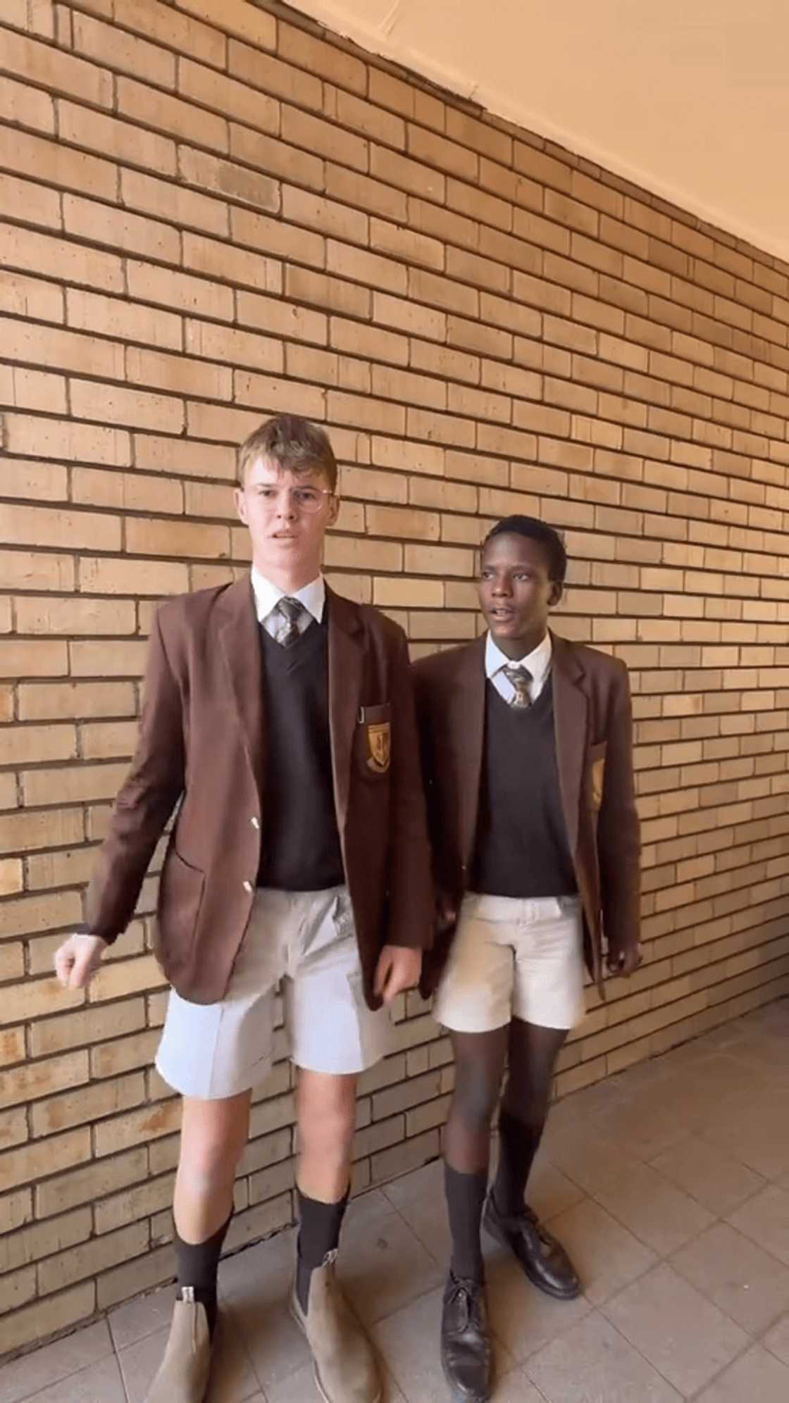 Two schoolboys in brown blazers, ties and white shorts stand side by side against a brick wall, about to start a dance, in a frame from a viral TikTok video. Two schoolboys in brown blazers, ties and white shorts stand side by side against a brick wall, about to start a dance, in a frame from a viral TikTok video.