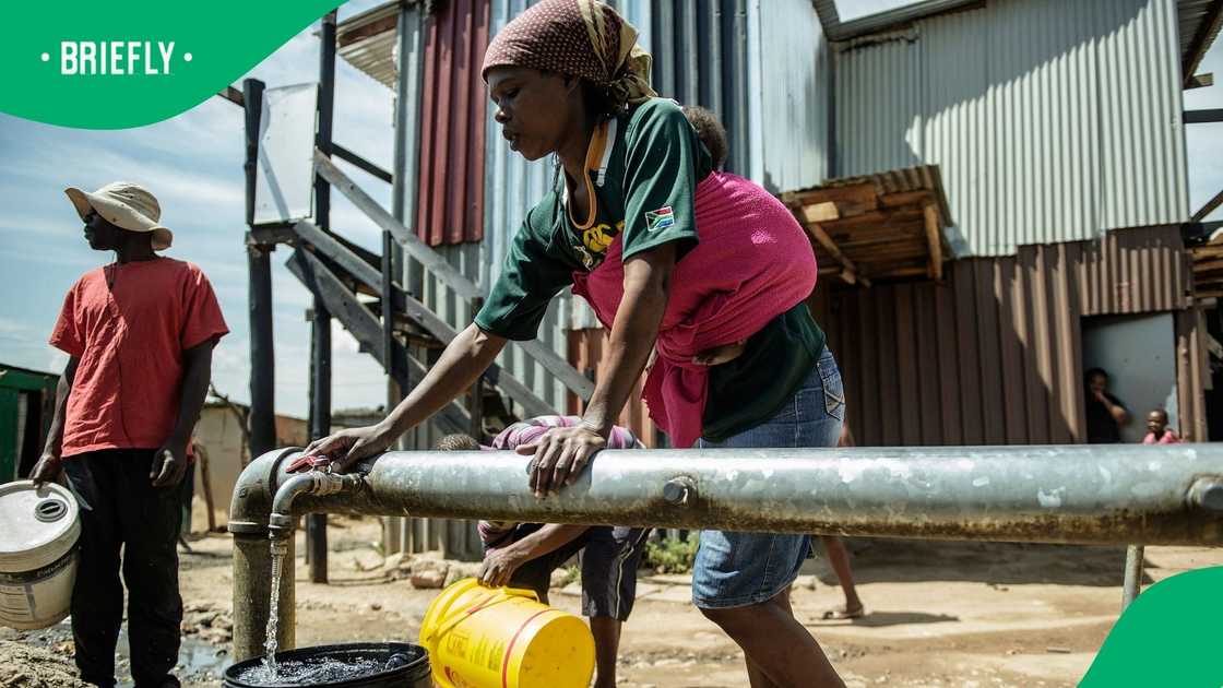 A South African woman with a baby wrapped in a pink blanket on her back turns a valve on a communal water pipe to fill a black bucket, while other residents wait with buckets in an informal settlement. A South African woman with a baby wrapped in a pink blanket on her back turns a valve on a communal water pipe to fill a black bucket, while other residents wait with buckets in an informal settlement.