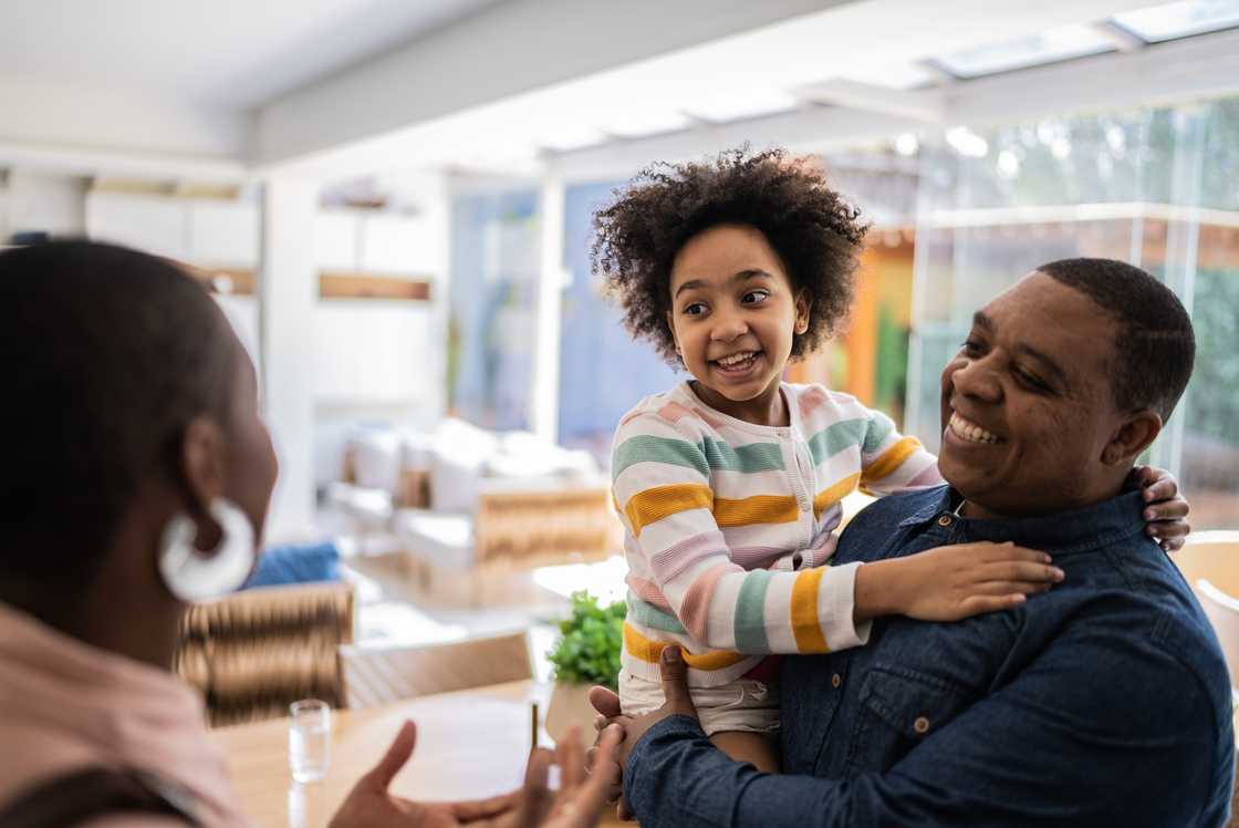 Smiling man holds a young girl in pastel stripes as they greet a woman indoors near large windows.
