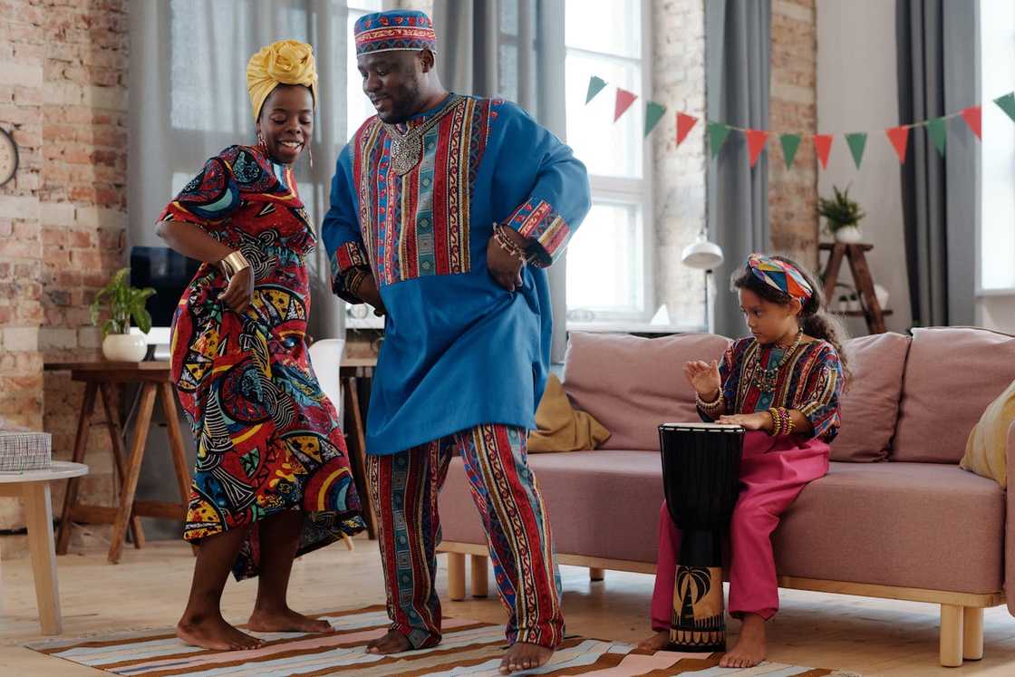 A family in colourful traditional clothing dances in a living room while a child plays a drum. A family in colourful traditional clothing dances in a living room while a child plays a drum.